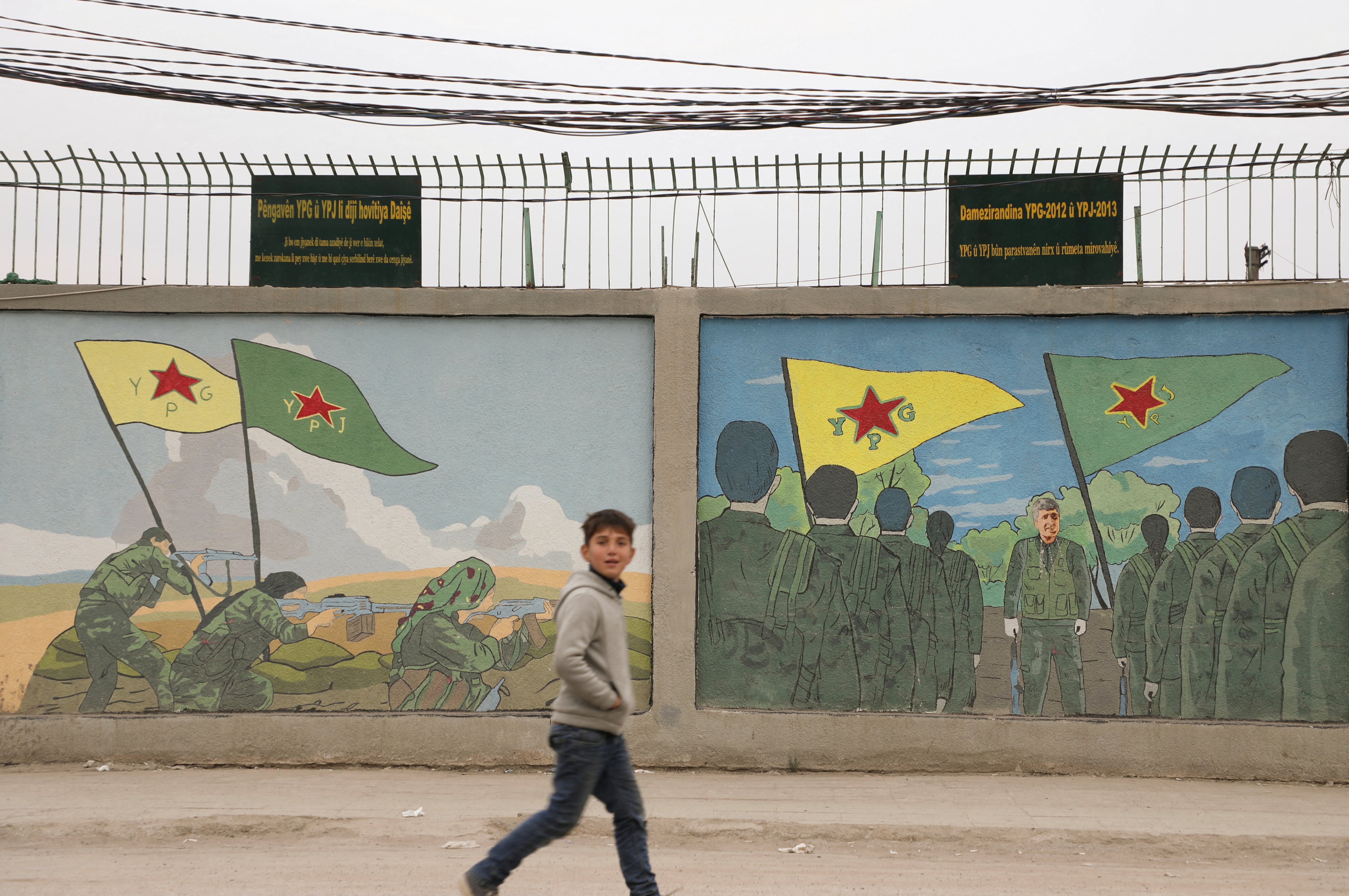 Un niño camina frente a murales con las banderas de las YPG e YPJ en Qamishli, diciembre de 2024. Semanas después, estas oficinas cerrarían sus puertas y retirarían los retratos de Öcalan mientras el mapa de Siria volvía a pintarse de un solo color tras 15 años de división. (REUTERS/Orhan Qereman/archivo)