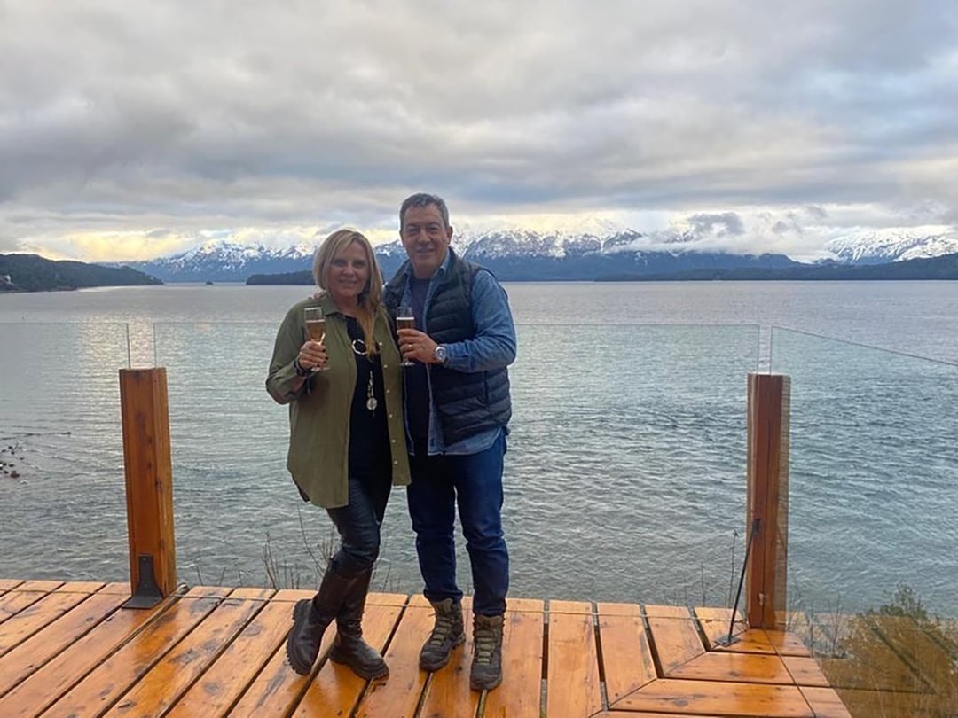 Chino Tapia y Viviana Juricka posan sonrientes en una terraza de madera con vista a un lago y montañas nevadas, celebrando con copas en mano.