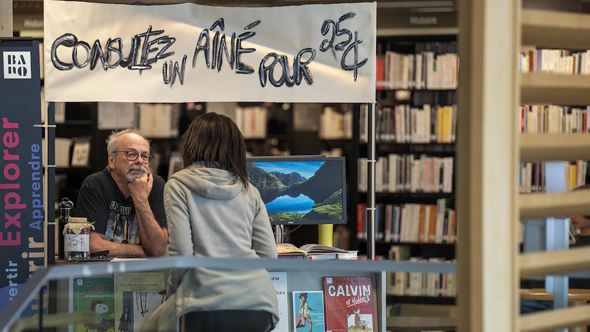 Hervé Simoneau, 77 años, responde preguntas desde su stand en la Biblioteca de Québec. Sobre su cabeza, un cartel: 