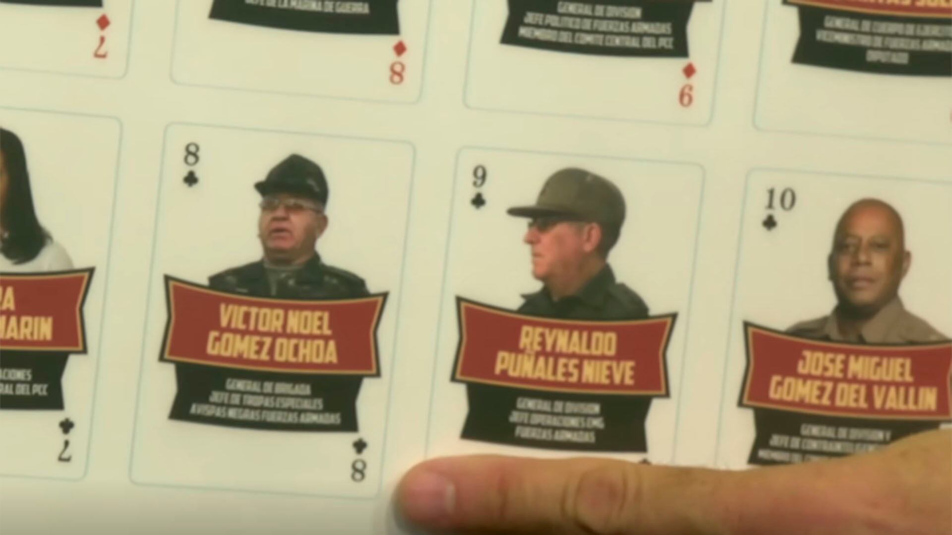 Una mano señala una baraja de naipes que representa a figuras de la dictadura cubana con sus fotos, nombres y rangos militares. (Captura de video)
