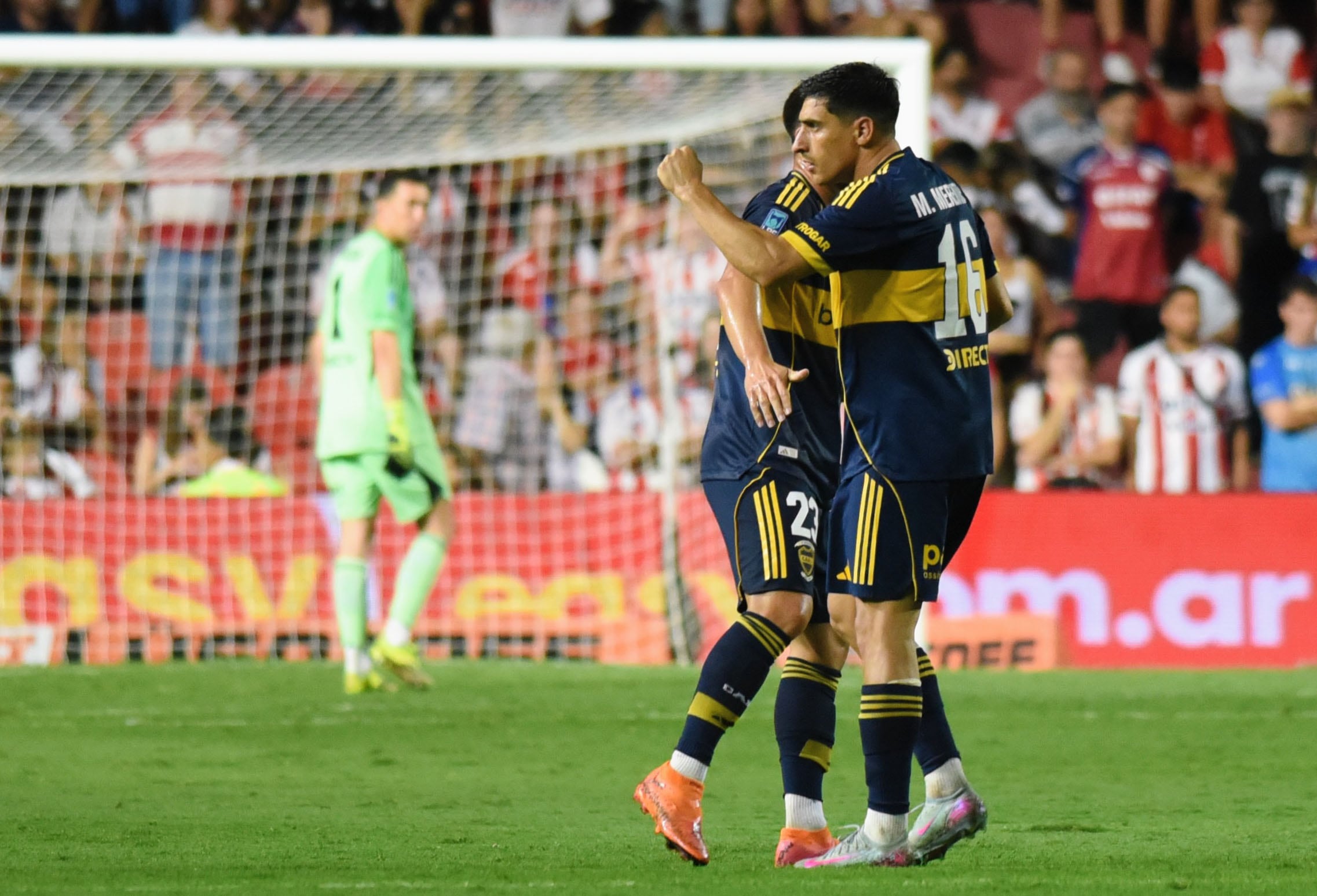 Dos jugadores de Boca Juniors se abrazan en el campo de juego, celebrando un gol, mientras el portero rival permanece en el fondo junto a la portería.