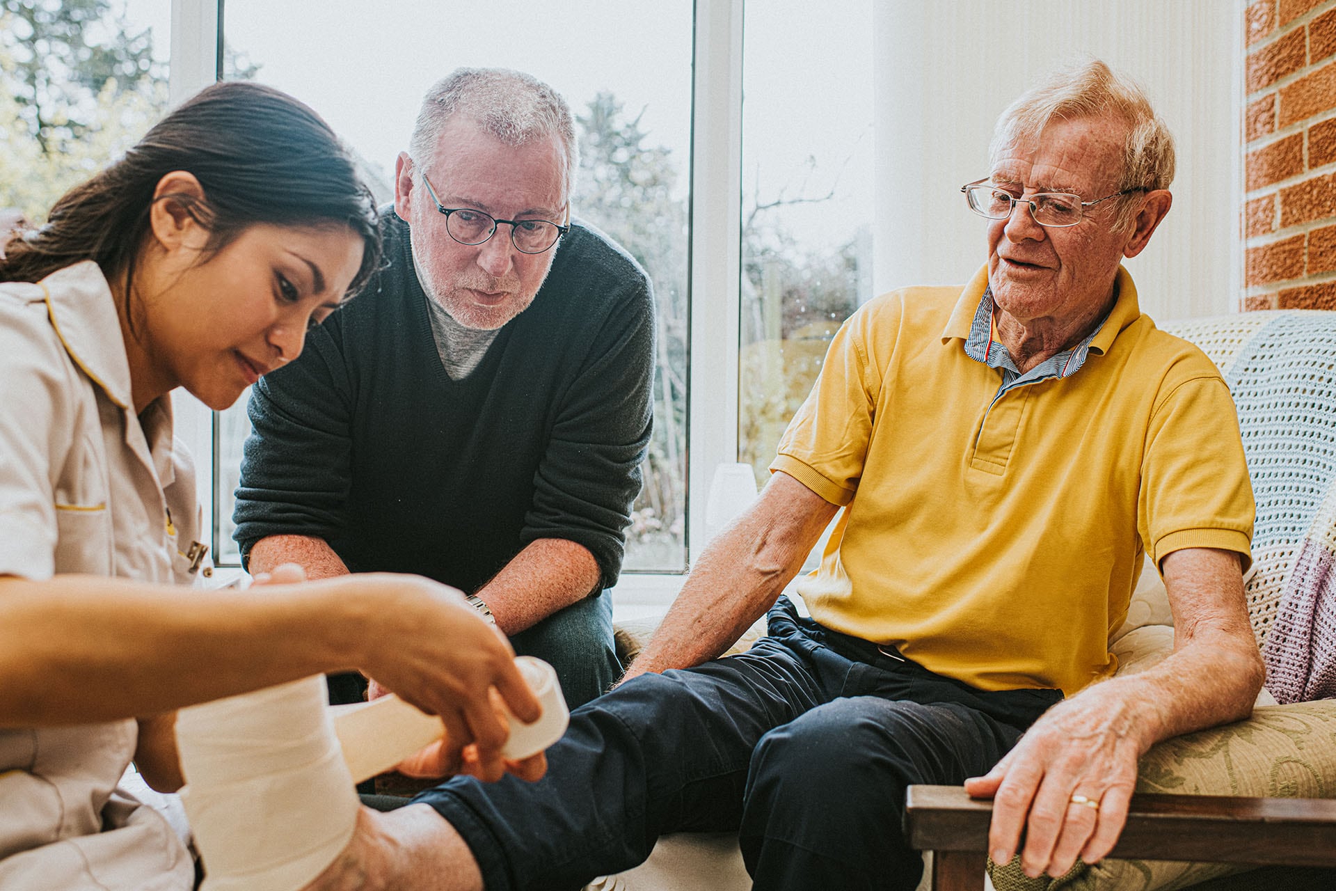 An elderly man seems shocked and in pain as a healthcare worker bandages his foot after a fall. An adult male watches over and looks concerned.