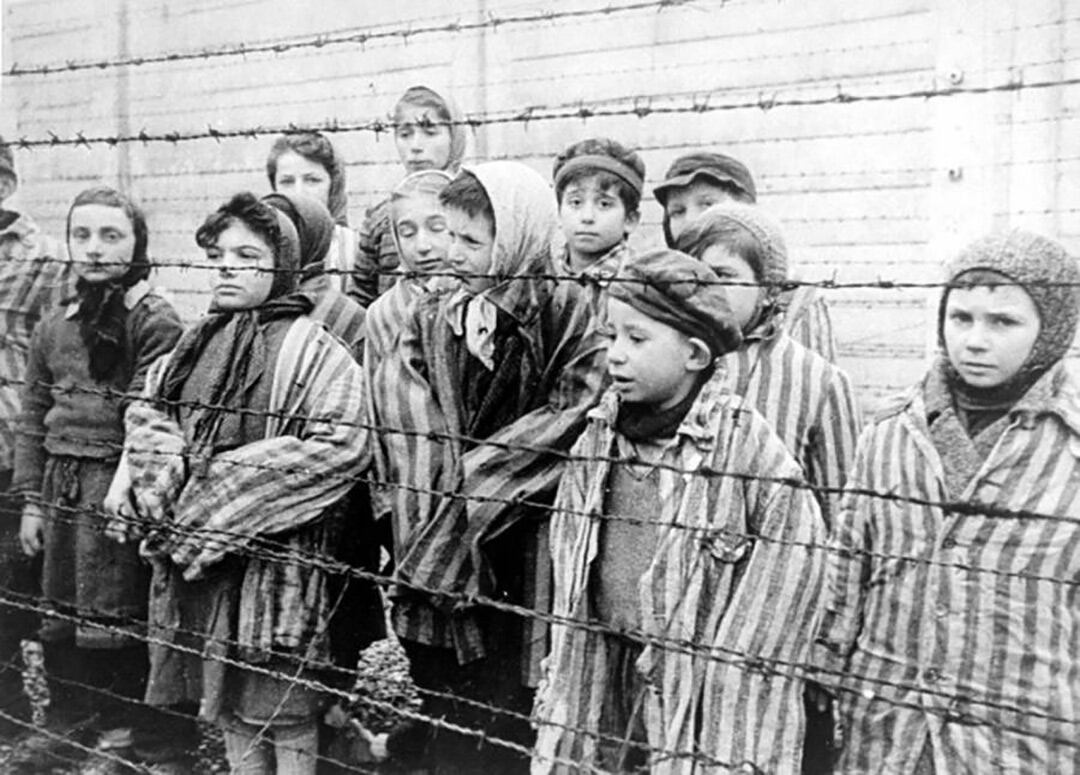 Un grupo de niños y jóvenes prisioneros, algunos con gorros, miran fijamente a través de alambradas en el campo de concentración de Auschwitz, reflejando el horror del Holocausto.