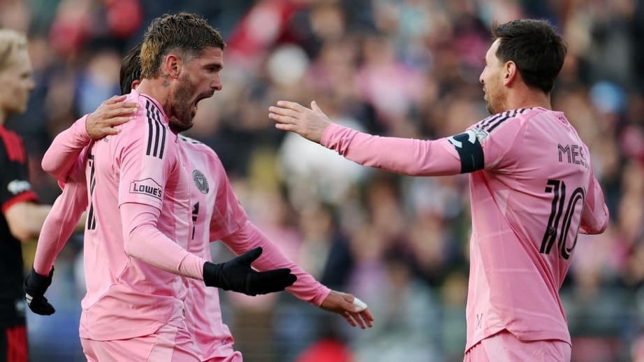 Lionel Messi y dos compañeros de equipo del Inter Miami celebran una jugada en el campo de juego de un estadio con aficionados de fondo.