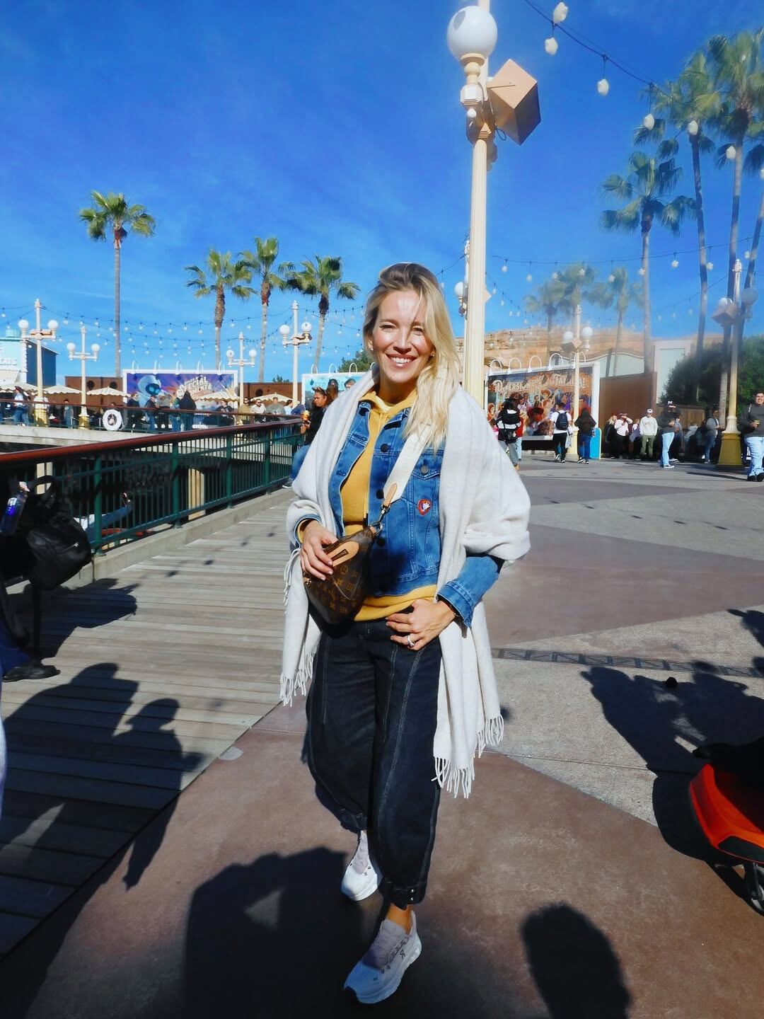 Luisana Lopilato sonríe ampliamente mientras posa al aire libre, con palmeras y edificios de un parque temático en el fondo, disfrutando de un día soleado.