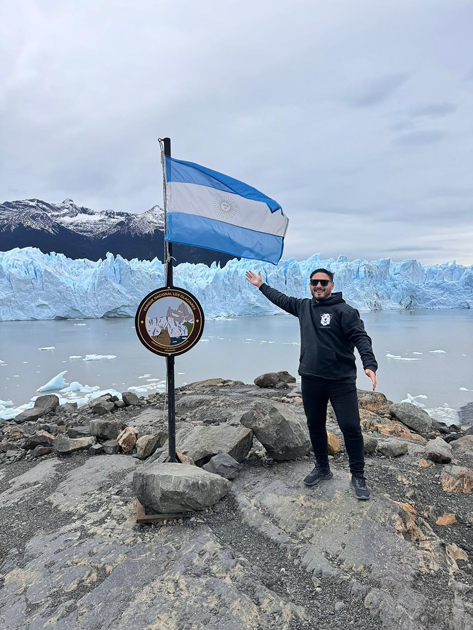 Hernán Lirio sonriente posa con la bandera argentina y un cartel del Parque Nacional Los Glaciares, con el imponente Glaciar Perito Moreno y témpanos flotantes de fondo.