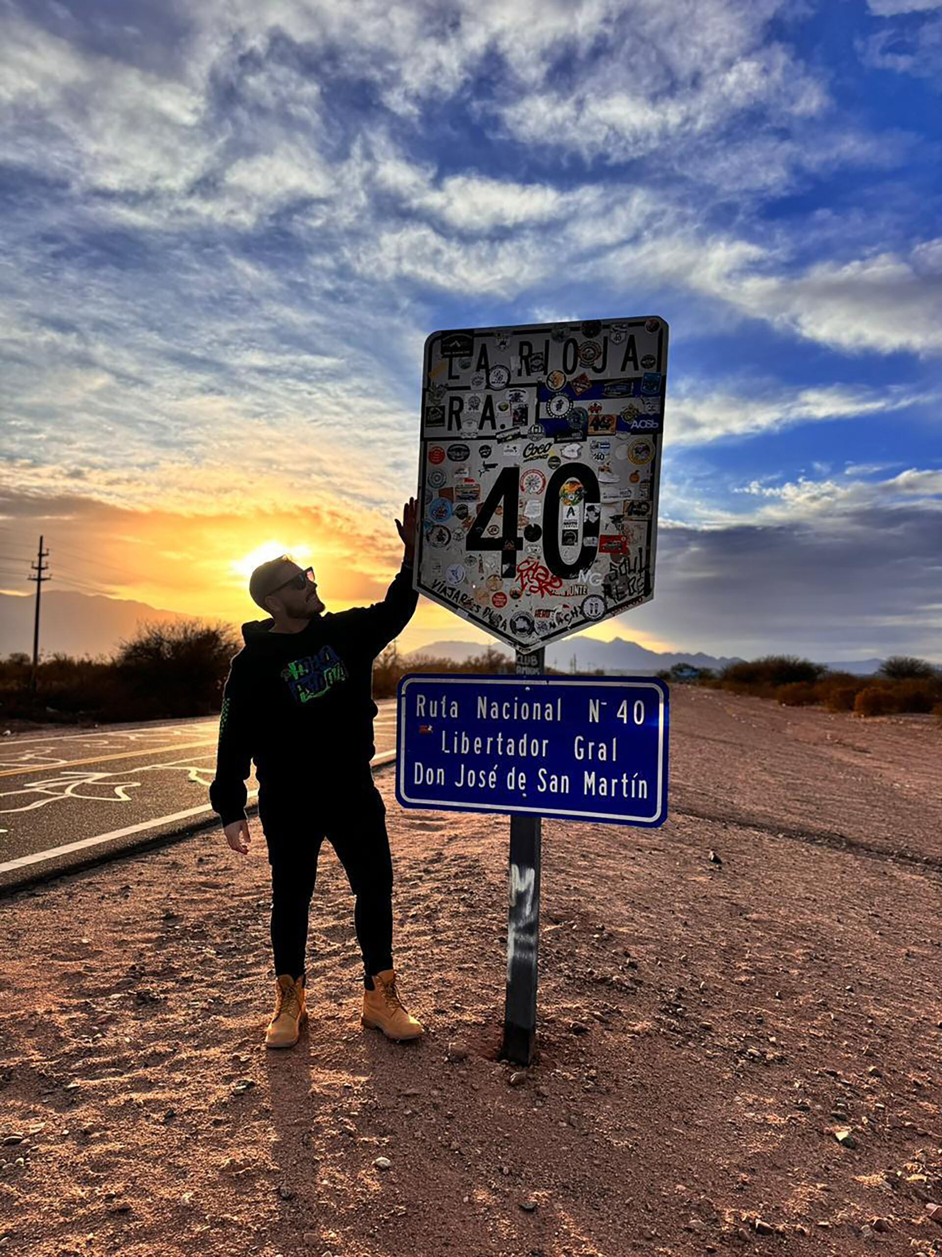 Hernán Lirio posa junto al emblemático cartel de la Ruta Nacional 40 en La Rioja, Argentina, al atardecer, con el cielo teñido de tonos cálidos.