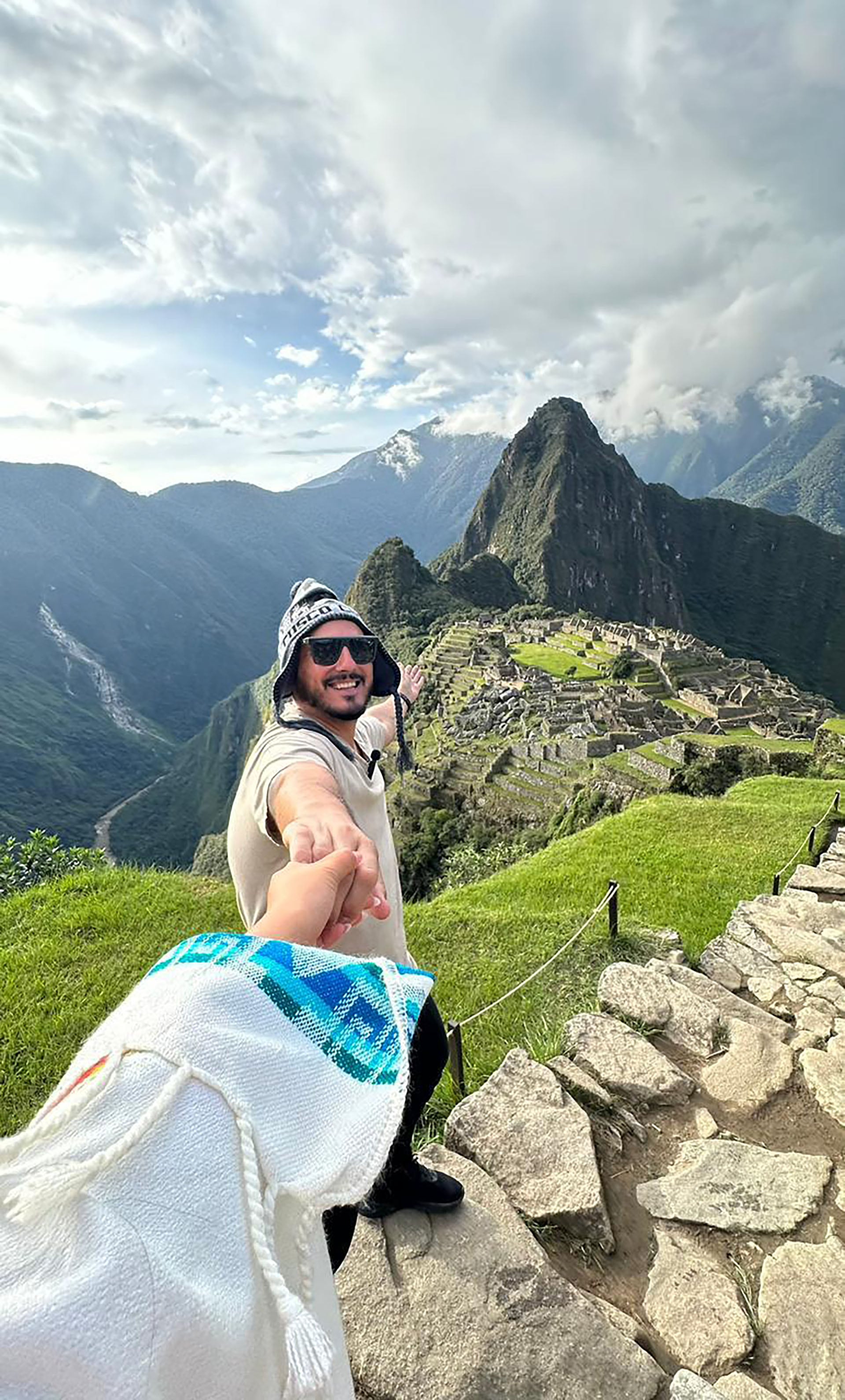 Hernán Lirio sonriente invita a la exploración, apuntando hacia la imponente ciudadela de Machu Picchu, enmarcada por montañas verdes y un cielo parcialmente nublado.