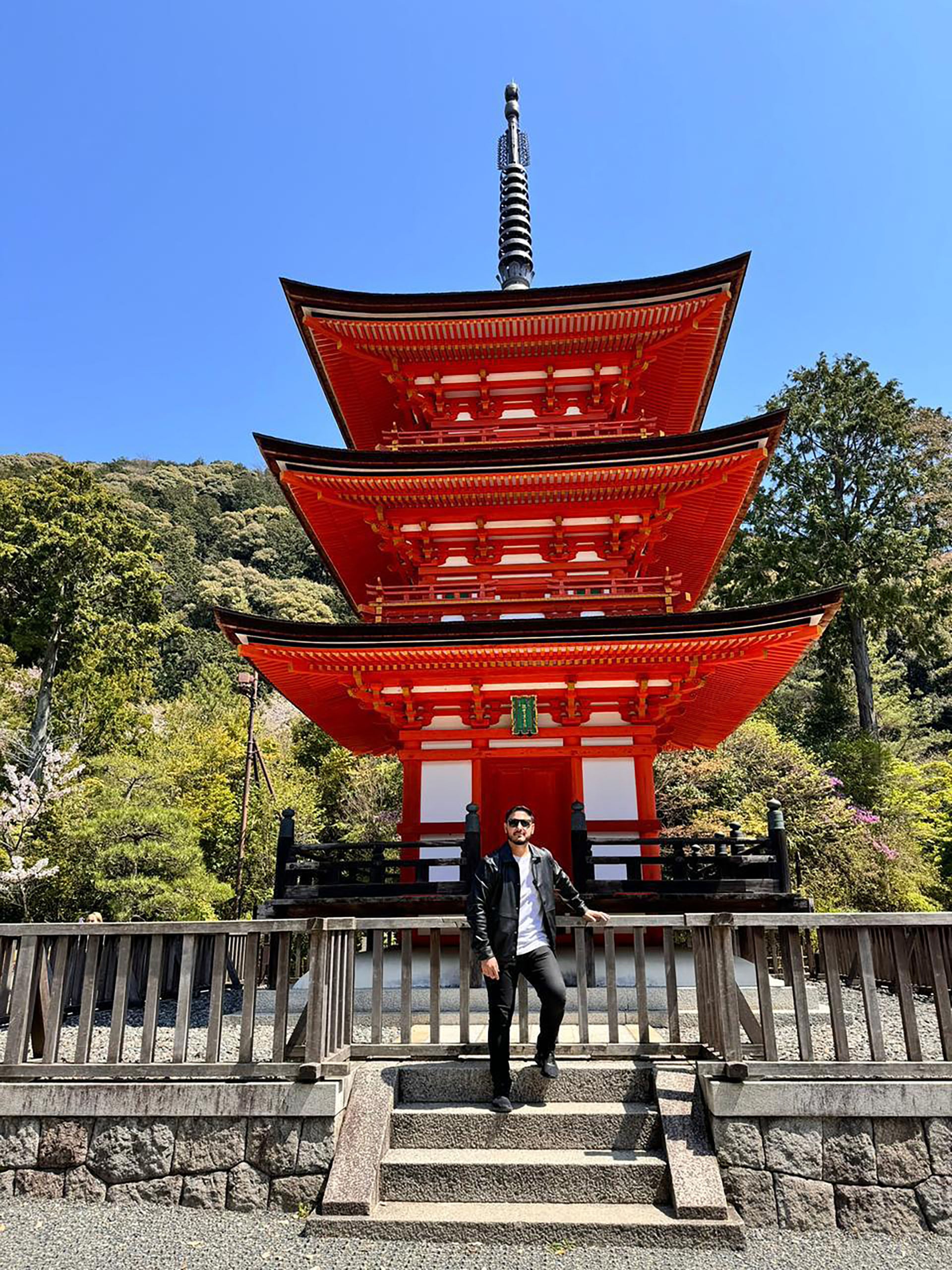 Hernán Lirio frente a una majestuosa pagoda roja de tres niveles en un santuario Shinto, enmarcada por exuberante vegetación bajo un cielo azul, celebrando la rica herencia arquitectónica.