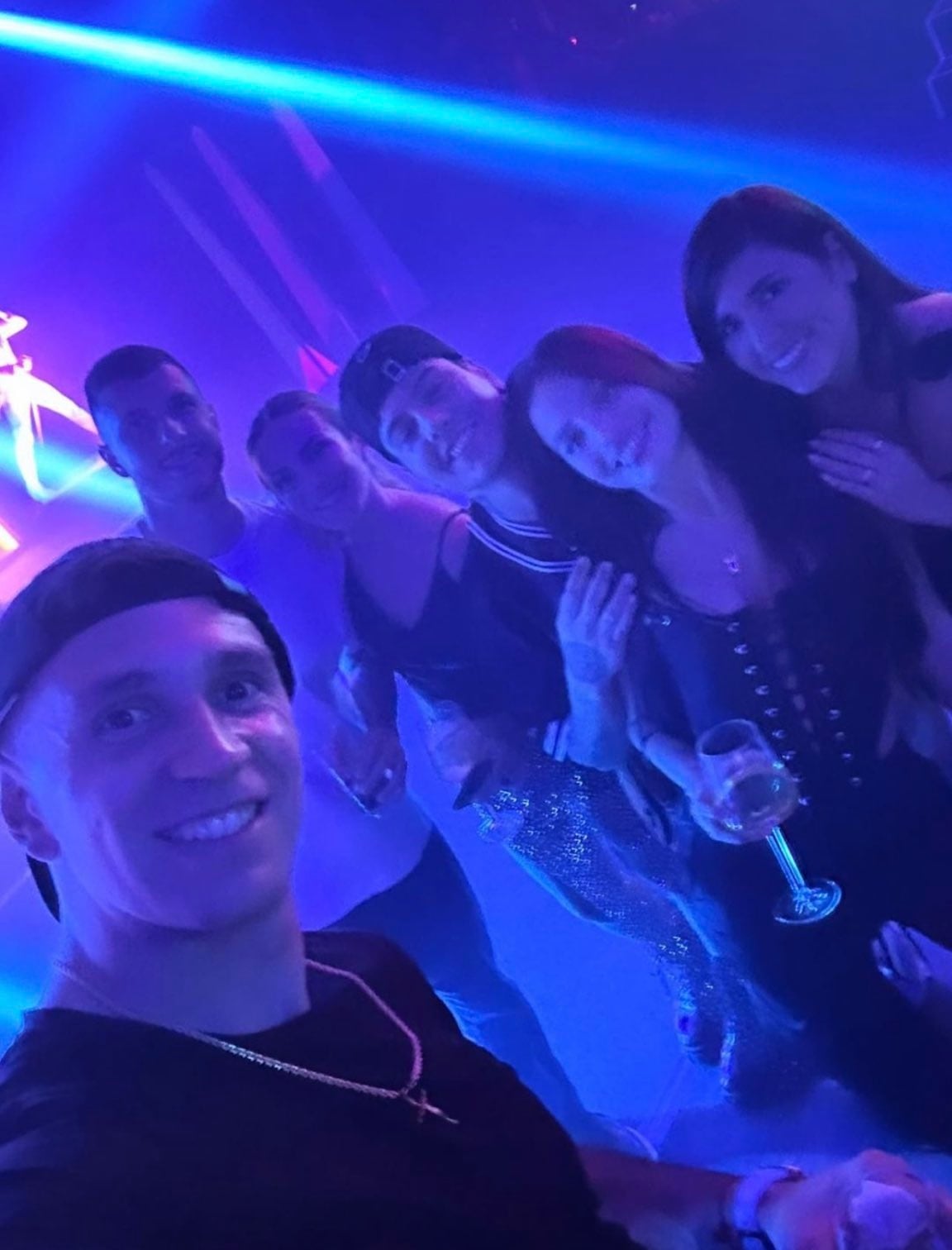Dibu Martínez, Emilia Mernes y Duki posan sonrientes junto a otros asistentes en la fiesta de la Selección Argentina, bajo luces azules vibrantes
