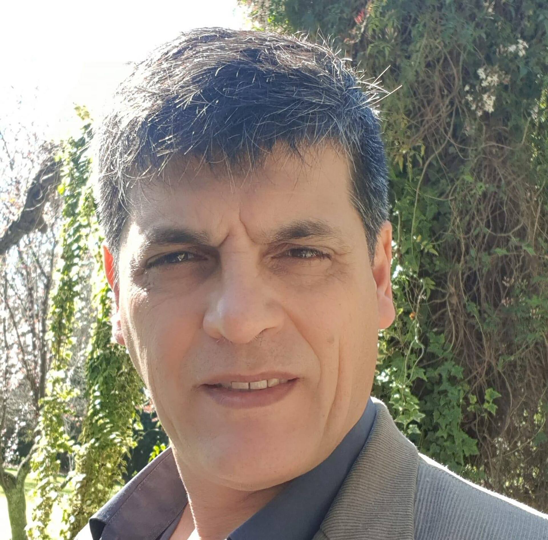 La víctima, un hombre de mediana edad sonríe levemente mientras posa para un retrato al aire libre con follaje verde y luz solar de fondo