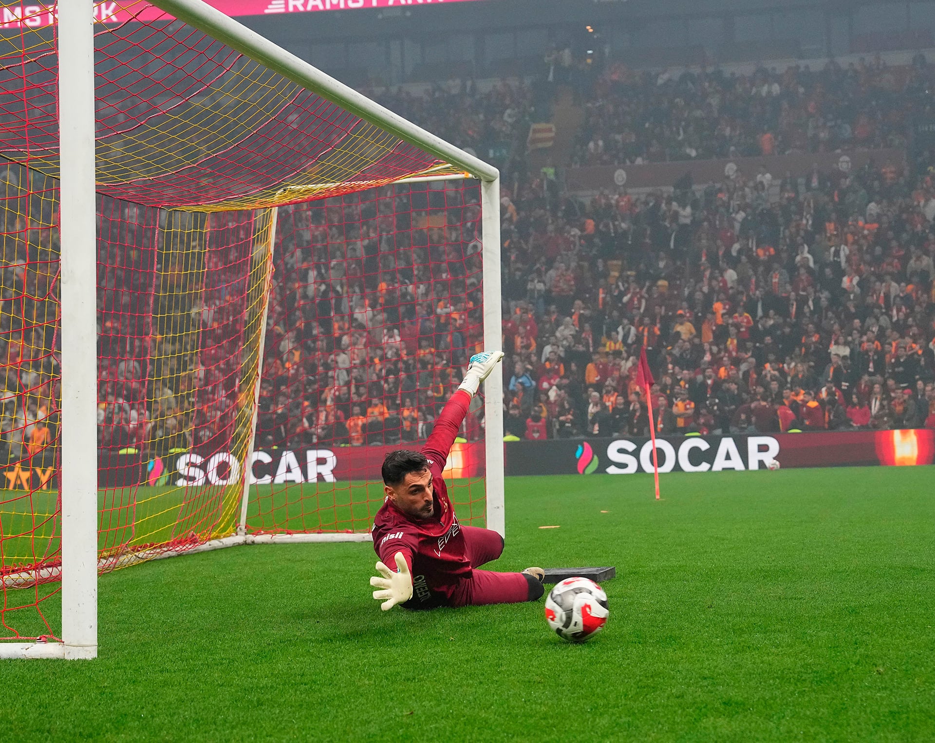 Un portero del Galatasaray realiza una estirada acrobática para atajar un balón durante un entrenamiento abierto, con el estadio lleno de aficionados presenciando la sesión. (@GalatasaraySK)