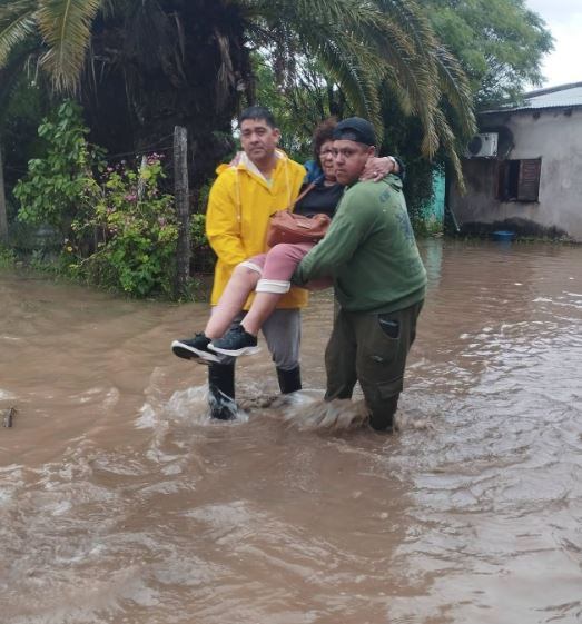 Dos hombres cargan a una mujer para ayudarla a cruzar una calle con graves inundaciones, en Los Juríes (Fm90.5 Santiago del Estero)