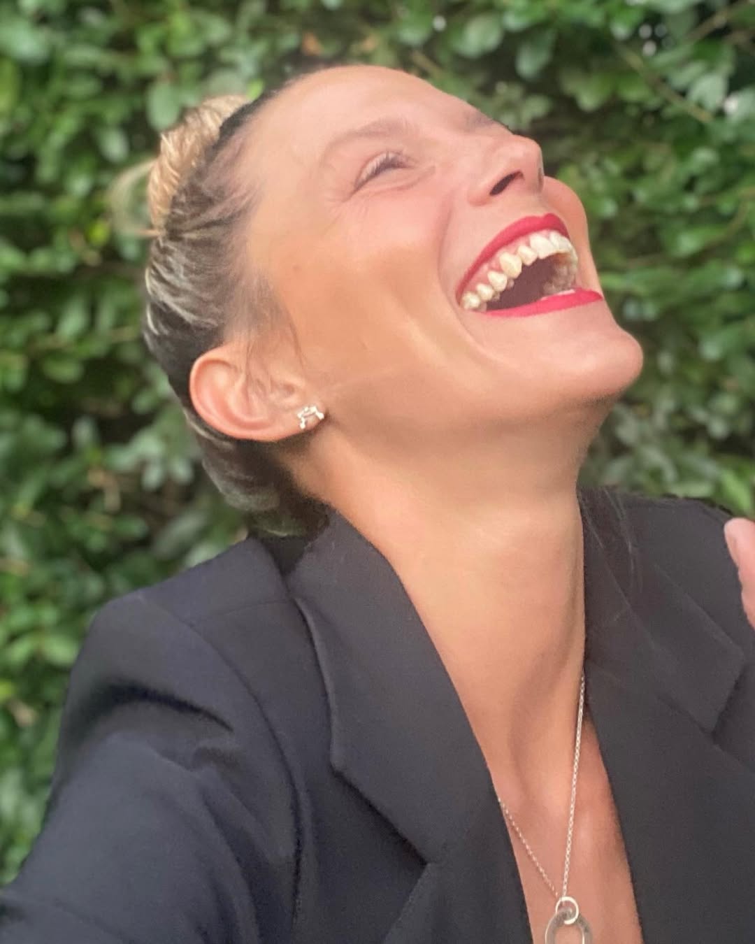 La reconocida actriz Manuela Pal se muestra feliz, riendo a carcajadas con el rostro hacia arriba y labios pintados de rojo, en un entorno natural.