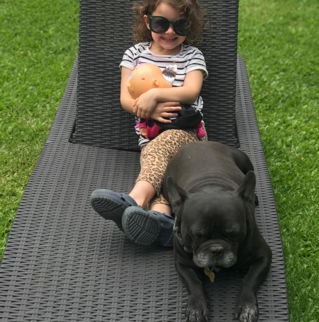 Filipa, sonriente con gafas de sol, abrazando una muñeca, comparte un relajante momento en una tumbona con Moro en un día soleado en el jardín 