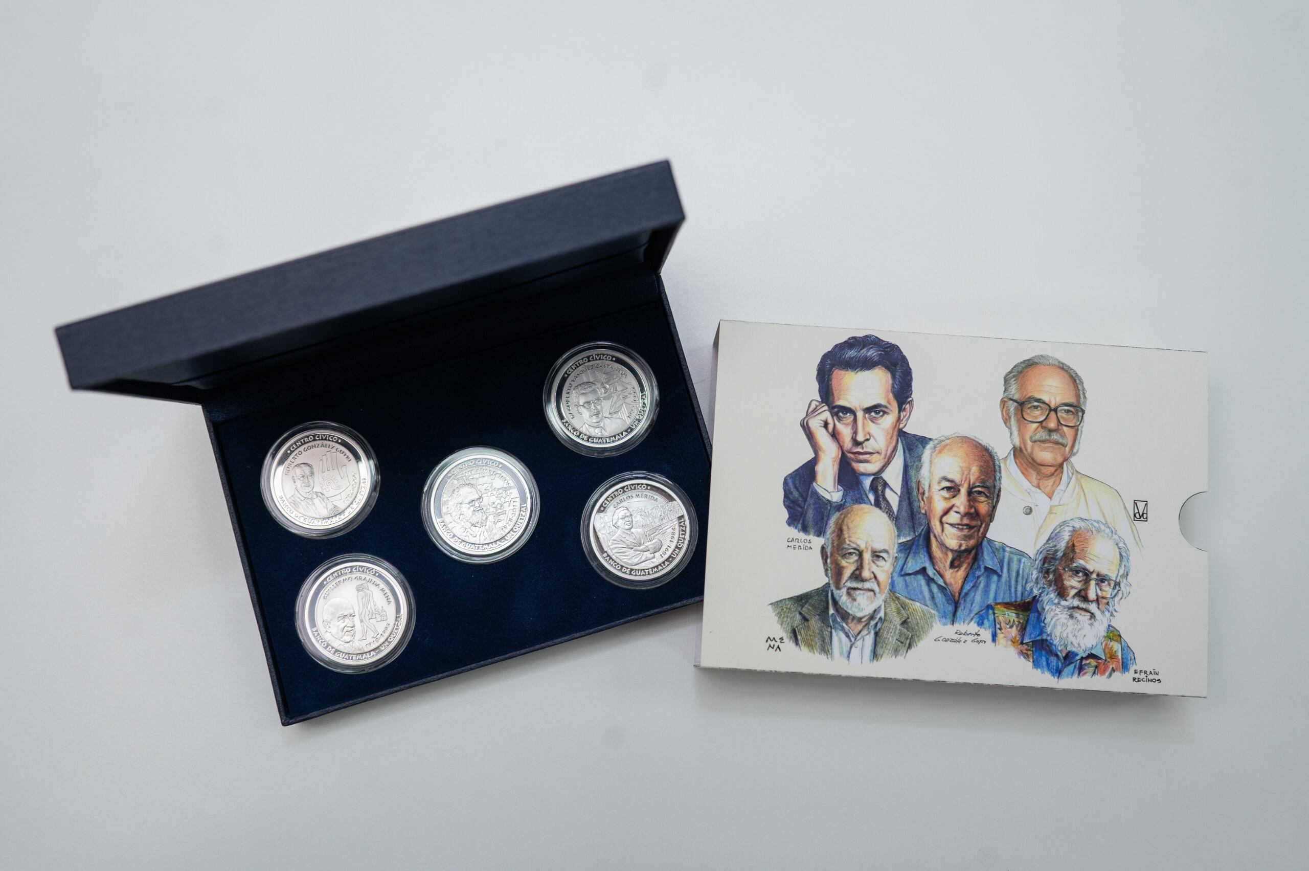 Una colección de cinco monedas conmemorativas del Banco de Guatemala, junto con un folleto ilustrado con los rostros de importantes figuras artísticas y culturales del país, se exhiben sobre una superficie blanca. (Banco de Guatemala)