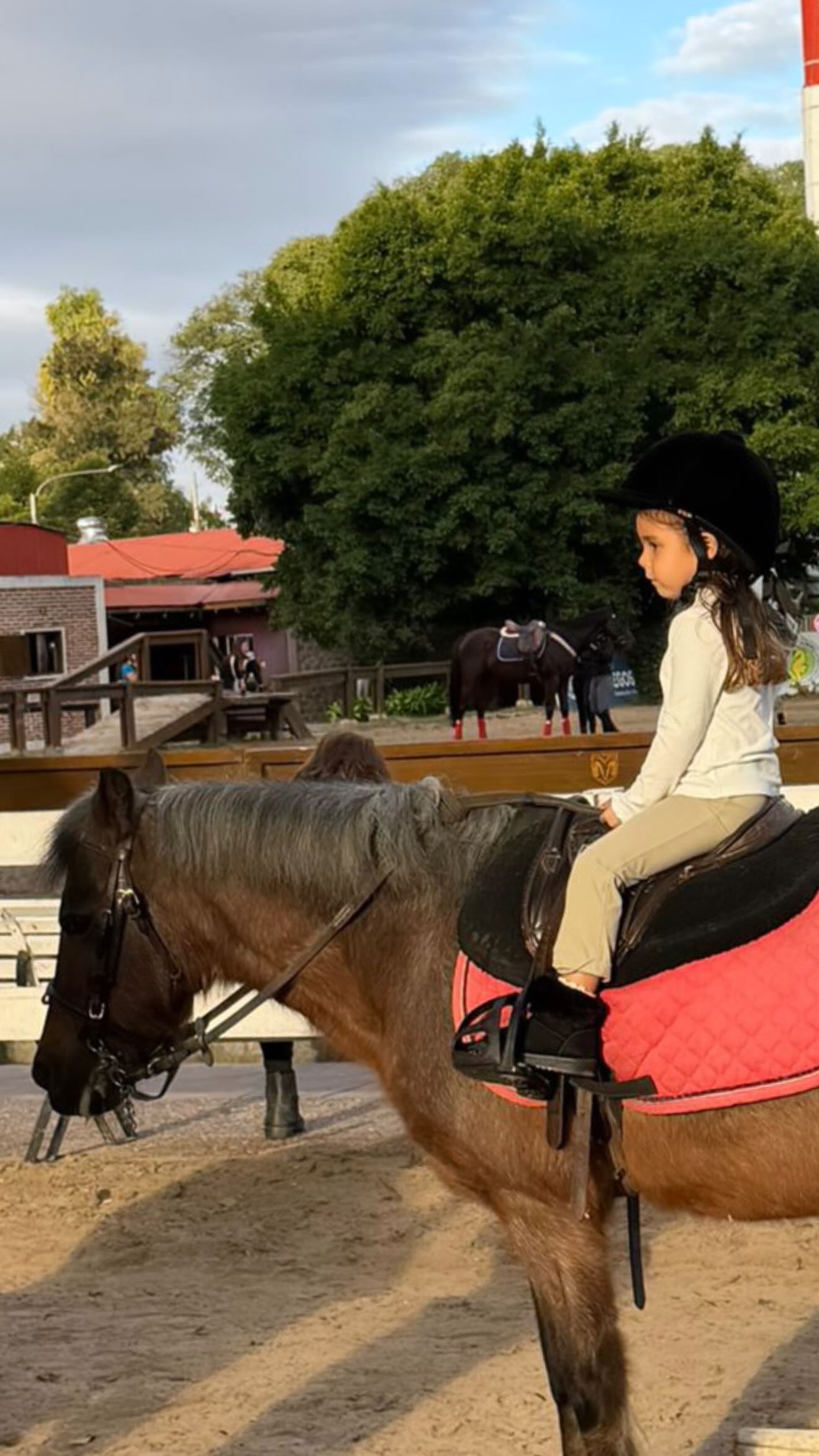 Sarah Burlando, junto a Ana García Moritán, participa con entusiasmo en su clase de equitación, montando un caballo en un picadero al aire libre.