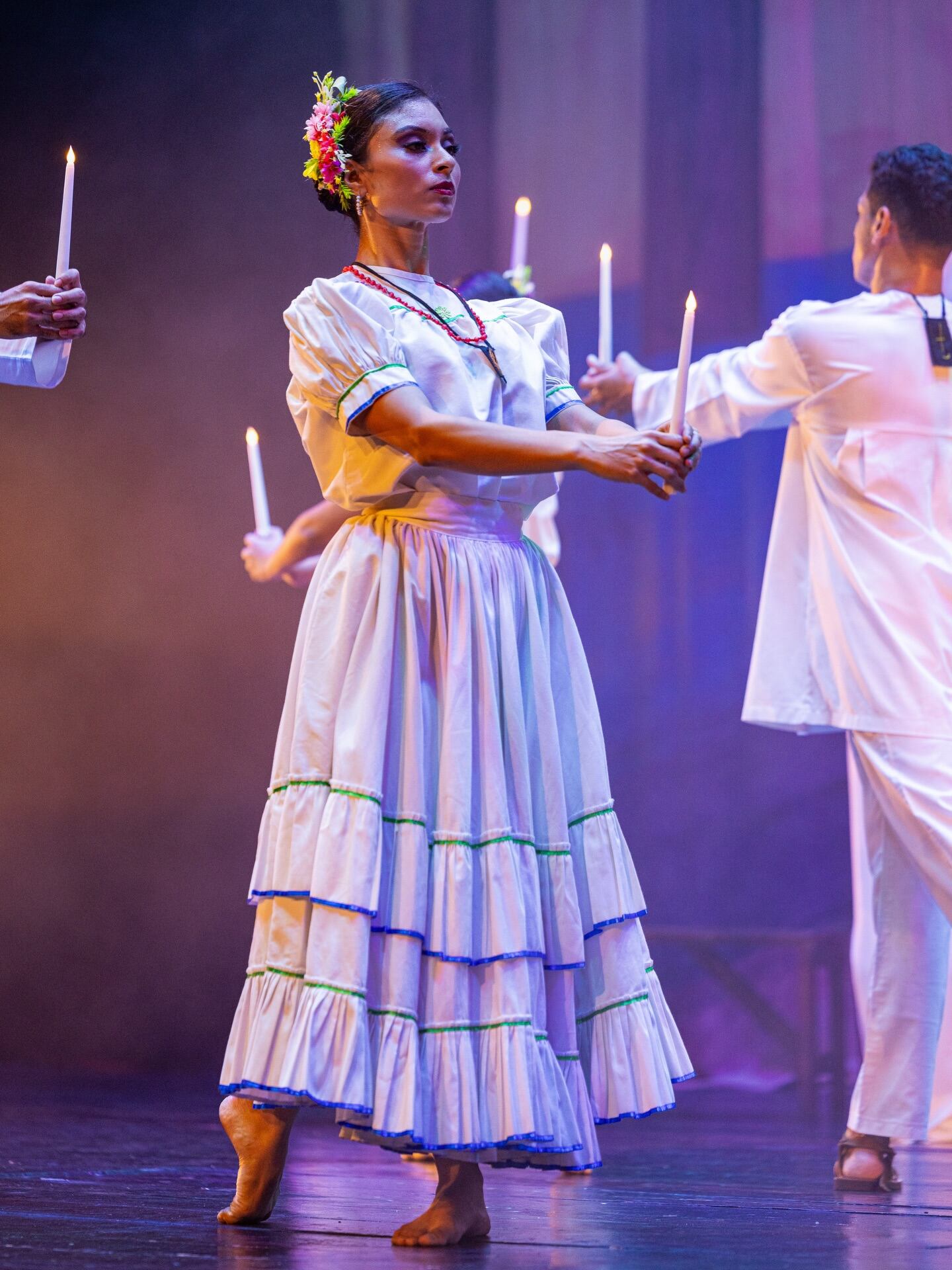 Una bailarina de danza folclórica, ataviada con un traje tradicional y flores en el cabello, realiza una coreografía solemne con una vela encendida, acompañada por otros bailarines en un escenario tenue. (Foto cortesía Ministerio de Gobernación)
