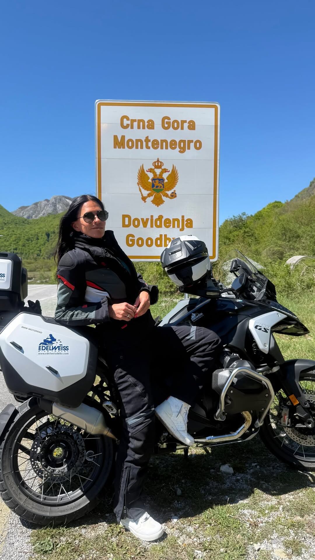 Carolina Baldini sonríe junto a su motocicleta BMW frente al cartel de bienvenida a Montenegro, iniciando una etapa más de su travesía de Croacia a Grecia en busca de libertad y nuevos comienzos.