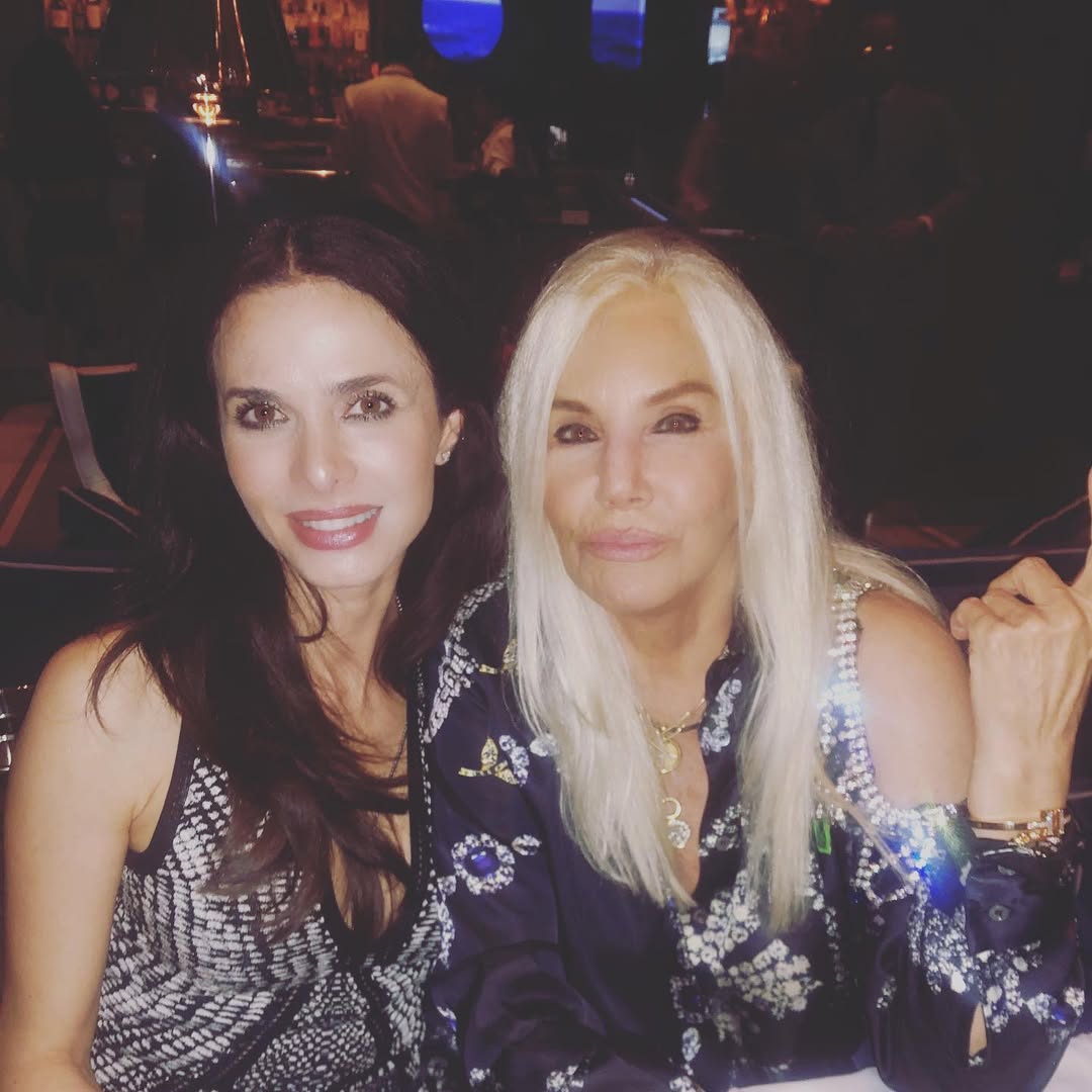 Rossana Almeyda, conocida por su vínculo con Marcelo Tinelli, posa sonriente junto a una amiga durante un evento social nocturno en un ambiente elegante.