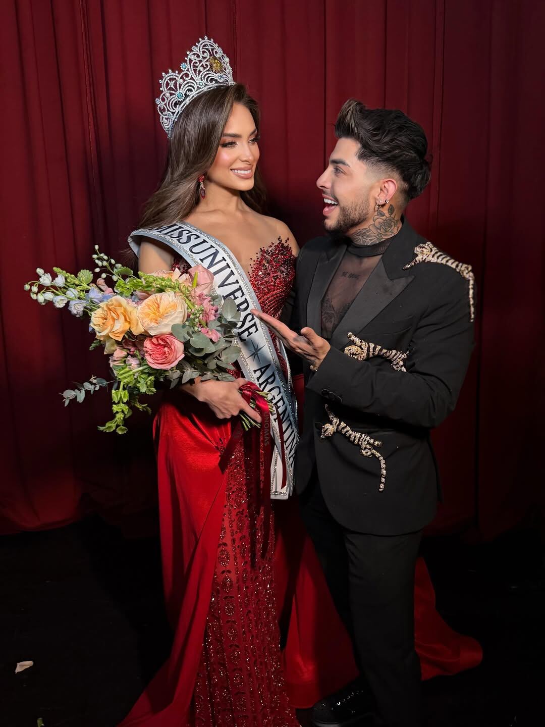 Tamara Rogouski, Miss Universo Argentina 2026, posa con su estilista Jorge Ramírez, luciendo su corona, banda y un ramo de flores, en un evento con fondo rojo.