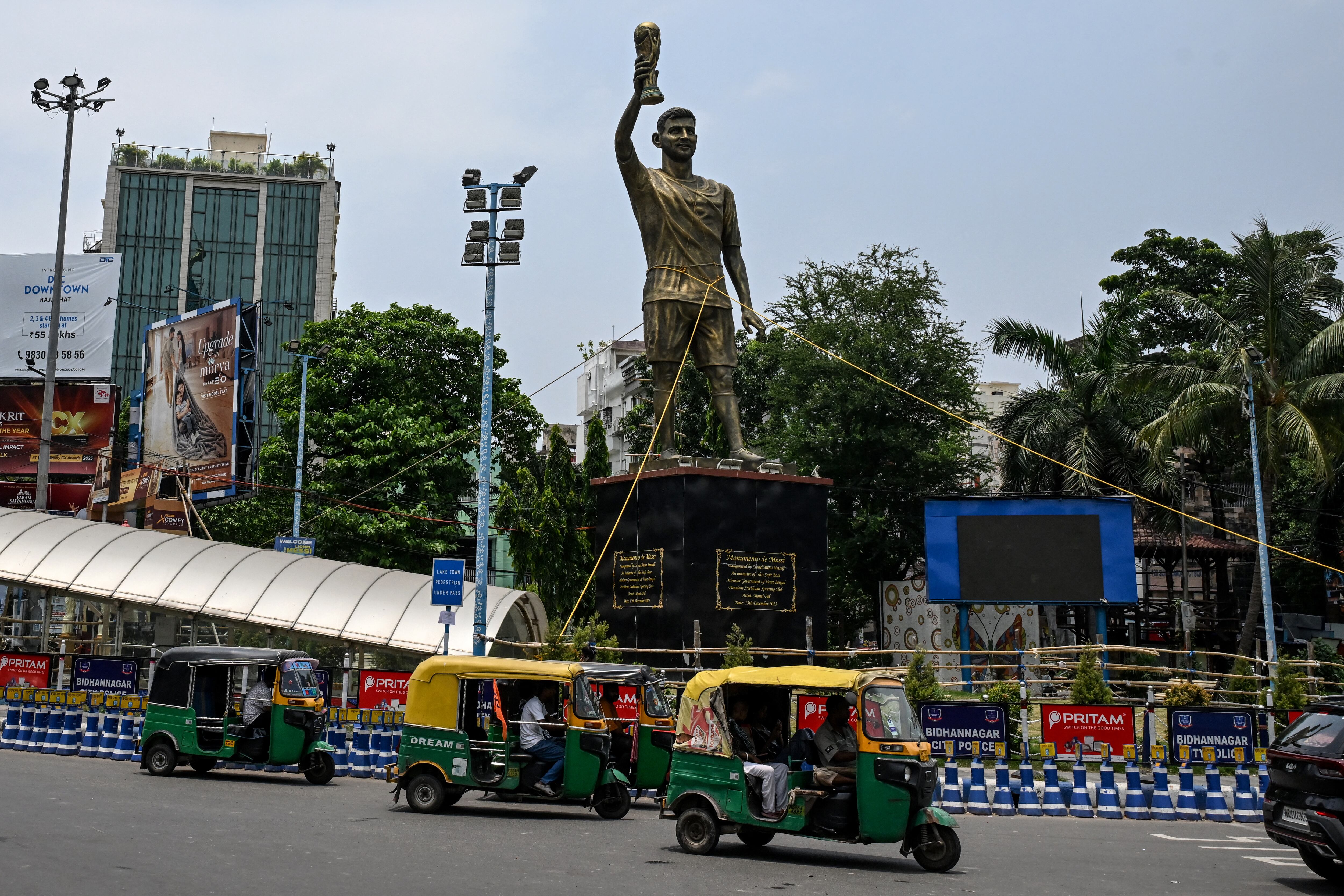 Los rickshaws pasan junto a una estatua del futbolista argentino Lionel Messi que ha sido atada con una cuerda para protegerla de la lluvia y las tormentas, antes de su reinstalación en un lugar más seguro, en Calcuta el 27 de mayo de 2026 (Foto de Dibyangshu SARKAR / AFP)