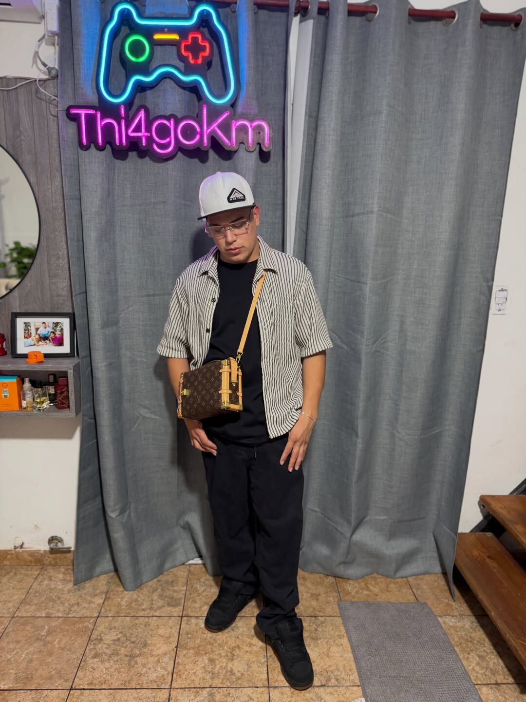 Thiago Medina posa con un atuendo casual que incluye una camisa a rayas, gorra blanca y un bolso cruzado de Louis Vuitton, destacando su estilo personal frente a un letrero de neón.