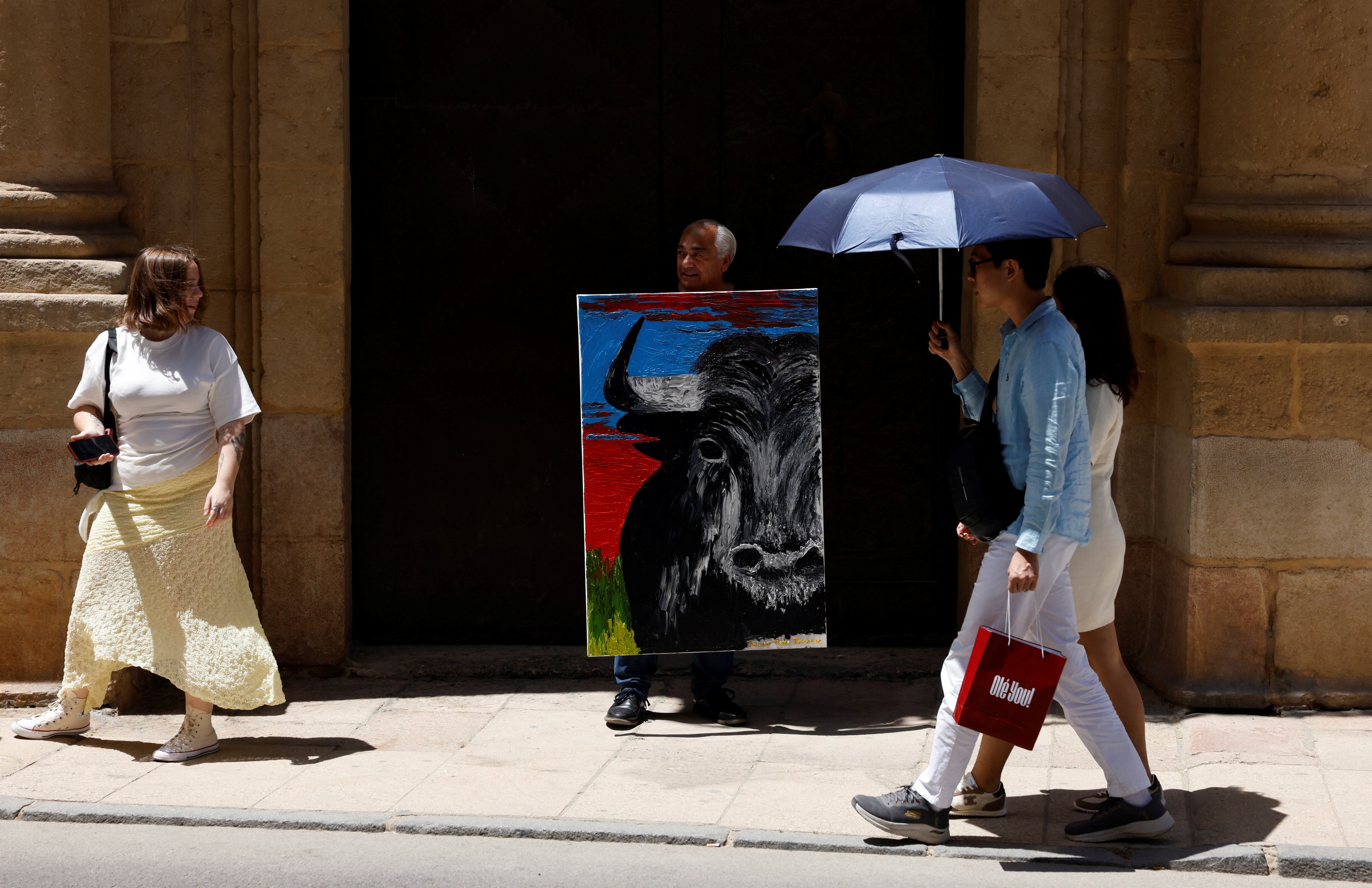 El artista español Javier Toro Truncer posa con su obra recién pintada frente a la plaza de toros de Ronda mientras turistas se protegen del sol con paraguas, en una jornada en que varias regiones de España batieron récords de temperatura para el mes de mayo. (REUTERS/Jon Nazca) 