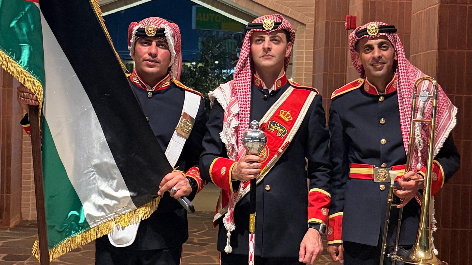 Tres integrantes de la banda militar de Jordania posan uniformados con un trombón, un bastón de mando y una bandera con franjas verde, blanca y negra.