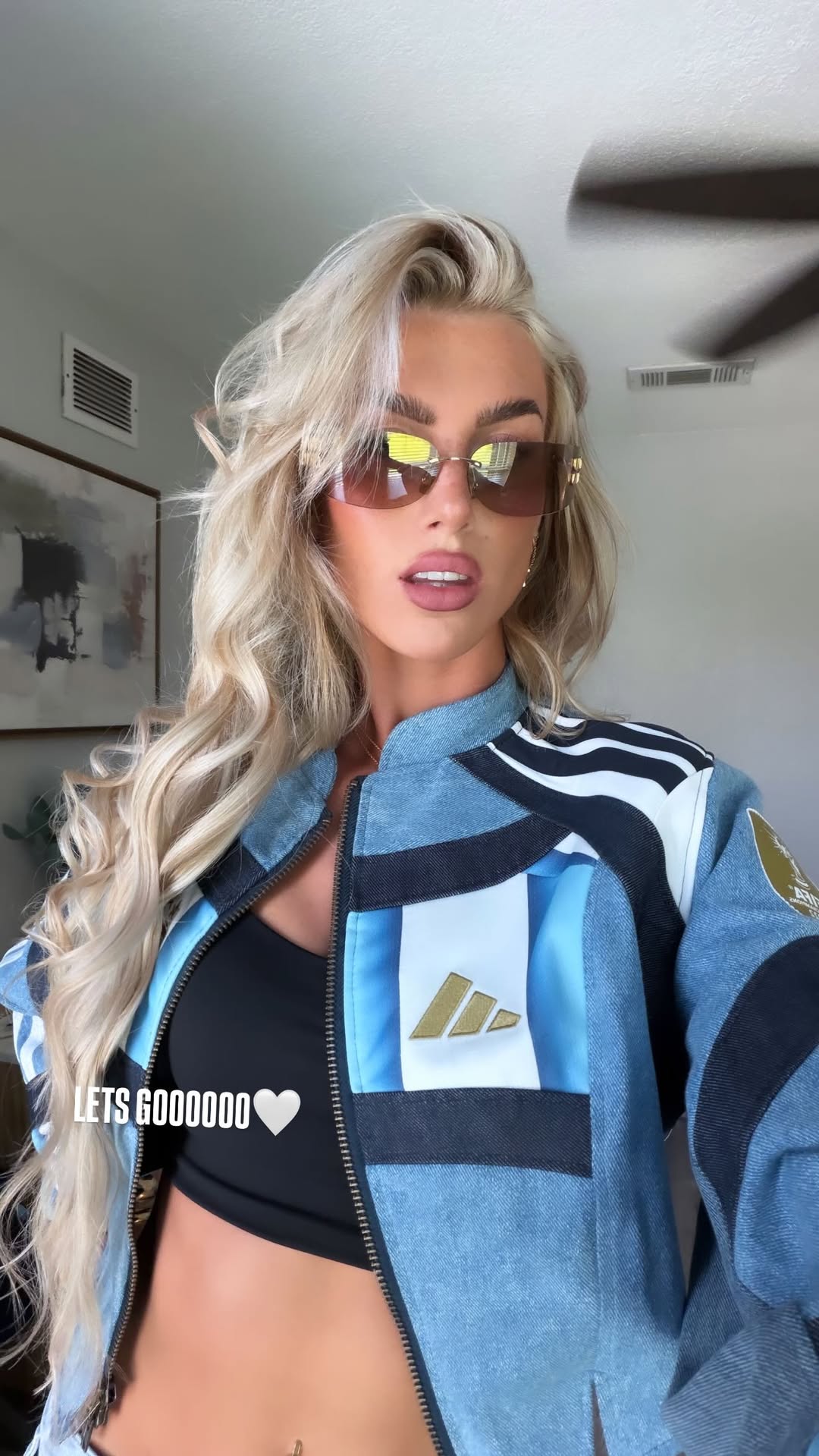 Kelci-Rose, pareja del futbolista Marcos Senesi, luce una chaqueta deportiva con los colores de Argentina, complementando su atuendo con gafas de sol y un top negro, en una imagen capturada en interiores.