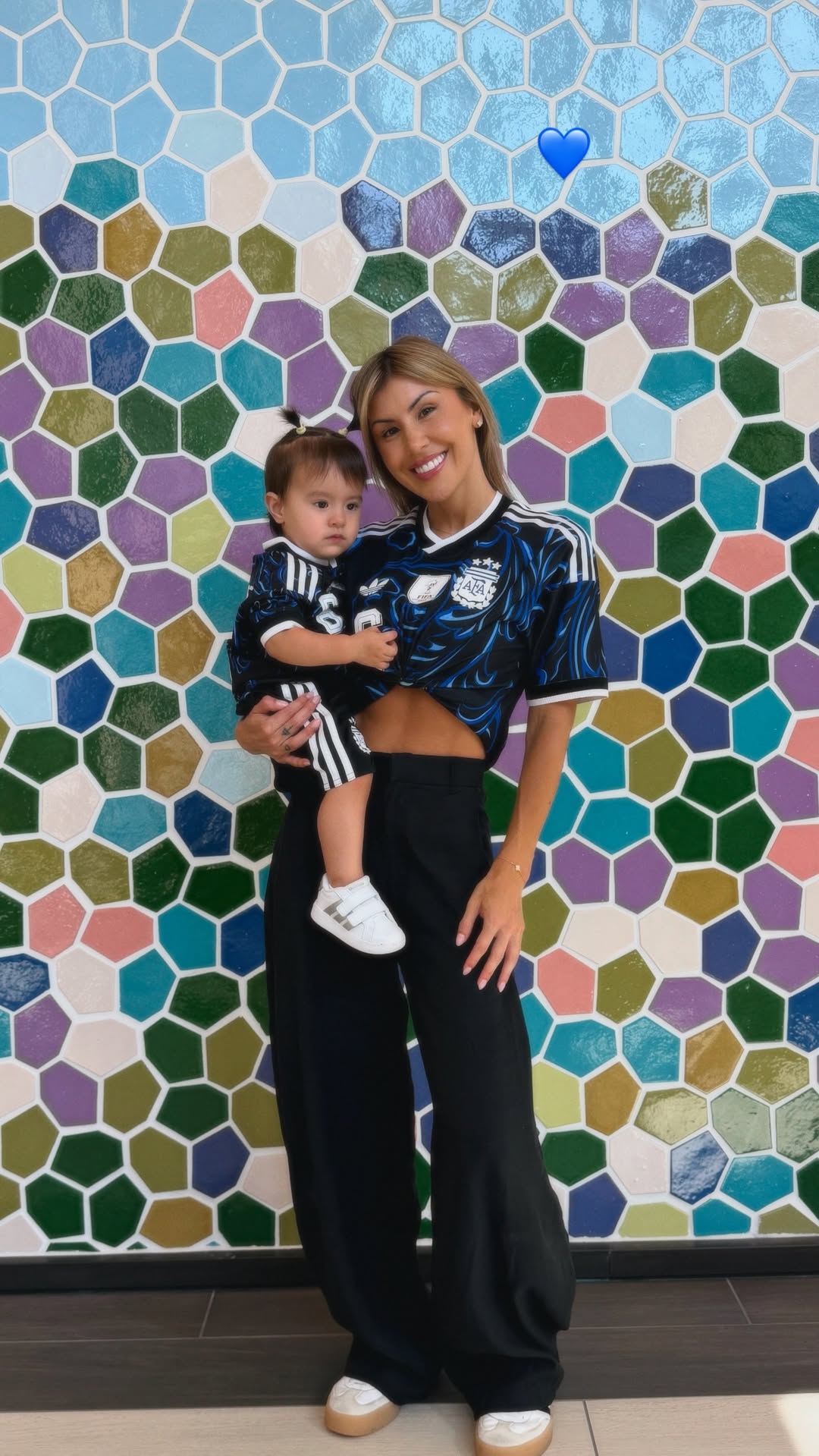 Muri López Benítez sostiene a su hija, ambas vestidas con la camiseta alternativa de la Selección Argentina, posando frente a una pared de mosaicos.