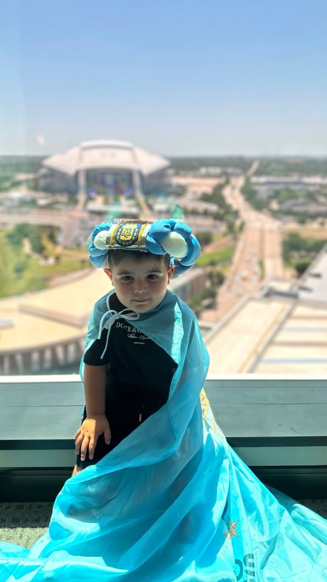 Benja Fernández, hijo del futbolista Enzo Fernández, posa con una capa celeste y blanca y un sombrero con el escudo de AFA frente al estadio de Arlington, sede del Mundial 2026.