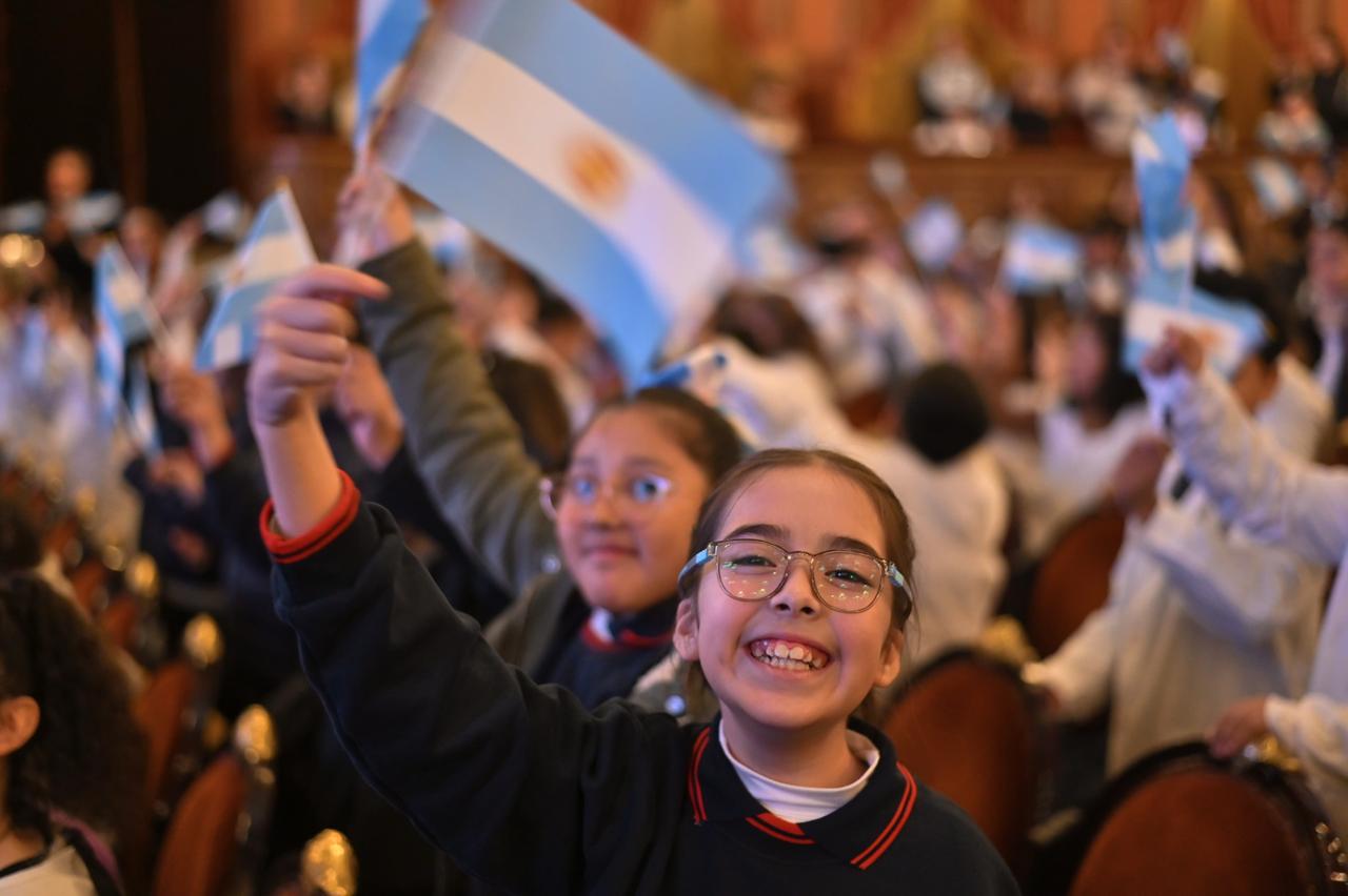 Una niña con gafas sonríe y levanta una bandera argentina entre una audiencia de niños que también agitan banderas en lo que parece un auditorio.