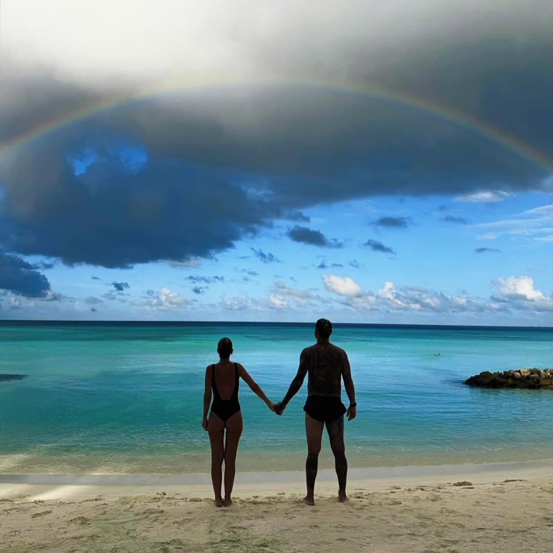 De espaldas y tomados de la mano, la pareja observó el mar desde la playa mientras un arcoíris cruzaba el cielo parcialmente nublado