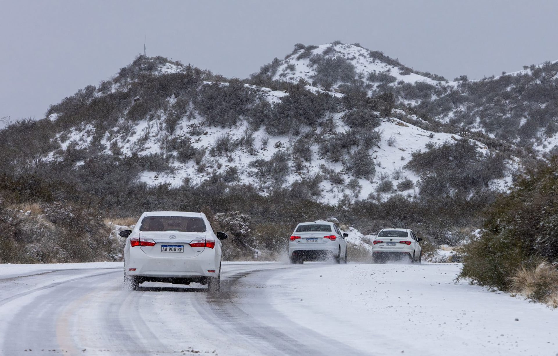 Varios vehículos circulan por una carretera cubierta de nieve en Malargüe, Mendoza, con montañas nevadas de fondo.