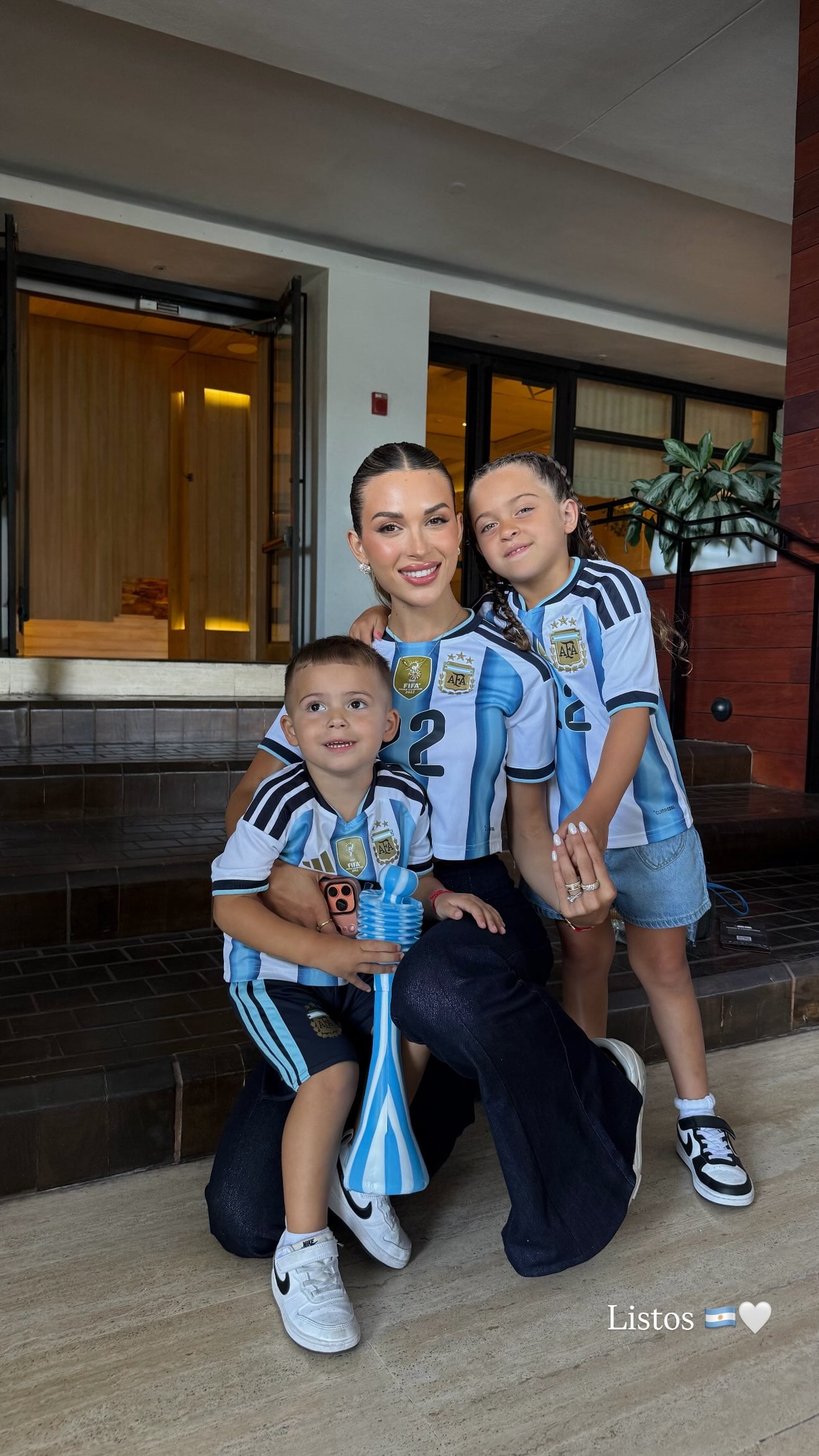 Agus Gandolfo, esposa de Lautaro Martínez, posa junto a sus dos hijos, quienes visten la camiseta de la selección argentina en un entorno interior.