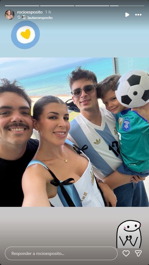 Rocío Espósito posa sonriente junto a Gerónimo Rulli, su hijo y otro hombre, quienes visten prendas deportivas de la selección argentina en un entorno de playa.