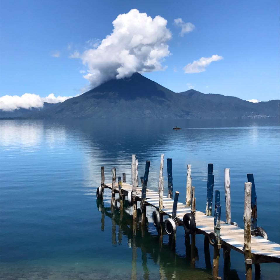 Un muelle de madera se adentra en las serenas aguas del Lago Atitlán, con un imponente volcán y nubes blancas reflejándose bajo un cielo azul en Santiago Atitlán, Guatemala. (página Lago de Atitlán, Guatemala)