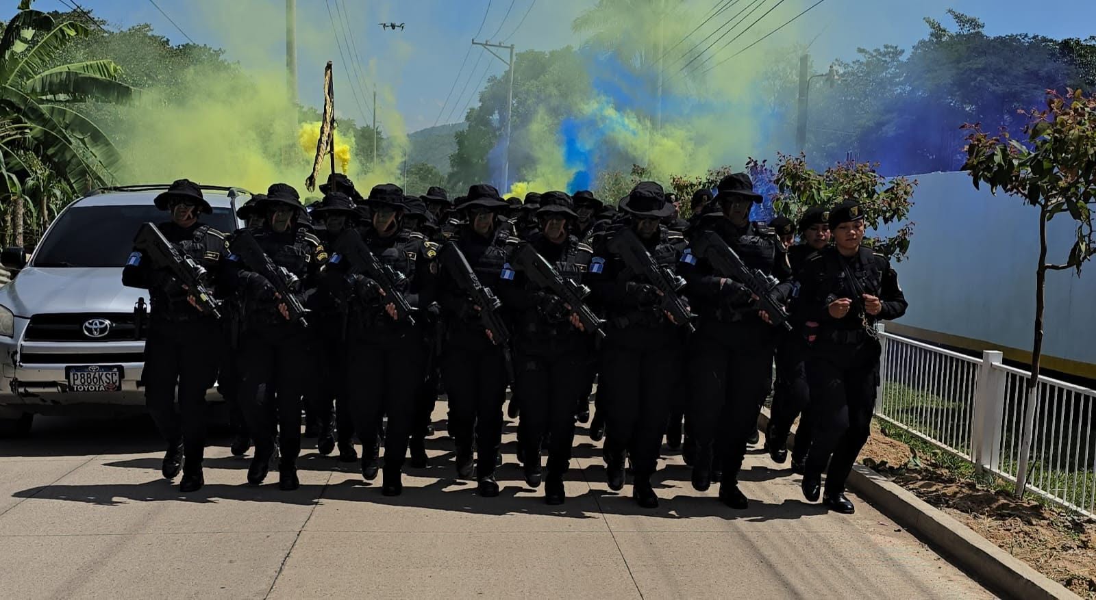 Agentes de seguridad de Guatemala marchan por una calle con uniformes negros y fusiles, acompañados de humo azul y amarillo, y un vehículo utilitario compacto. (Ministerio de Gobernación)