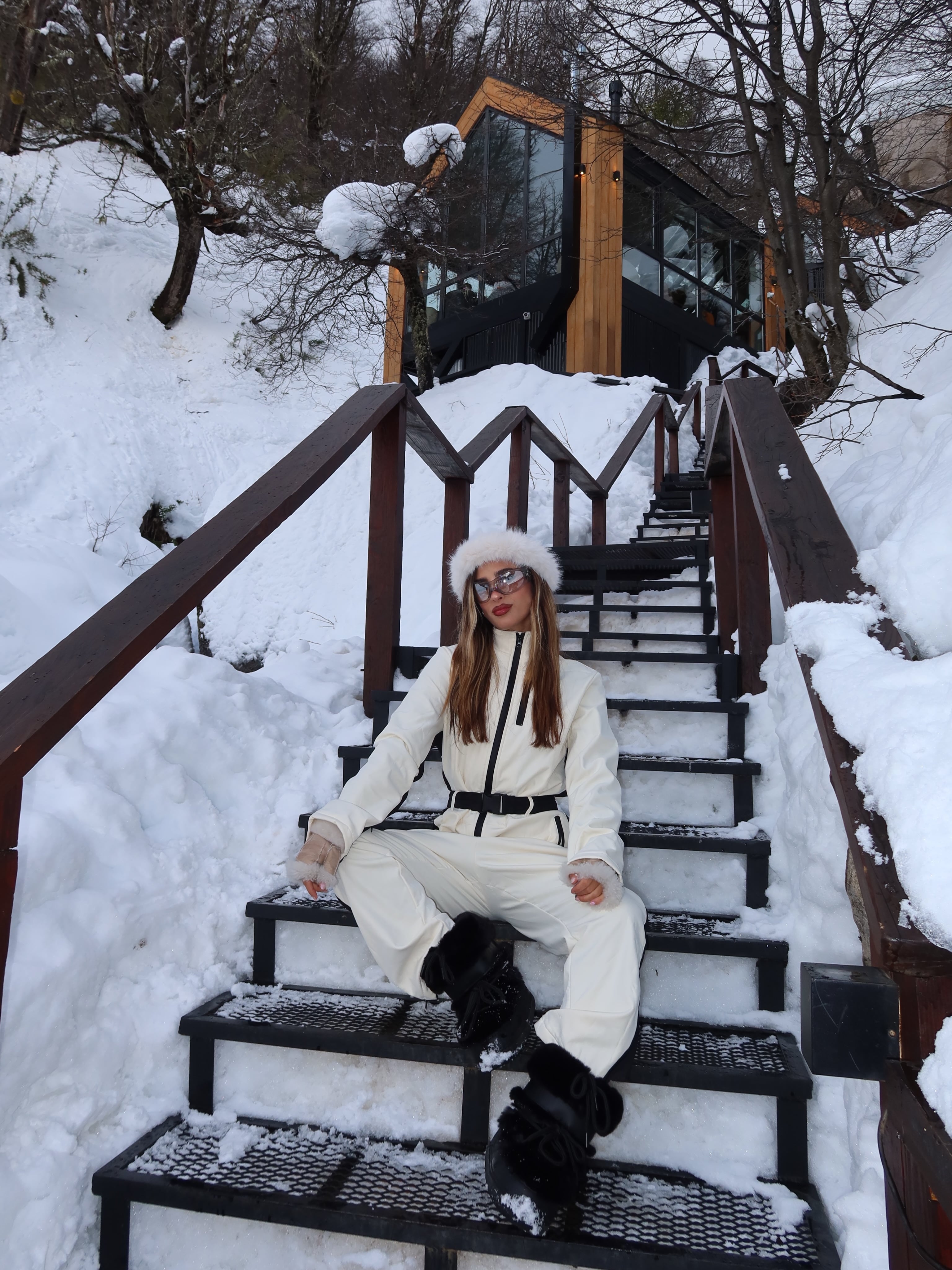 Julieta Poggio, sentada sobre los escalones nevados de la villa, con mameluco blanco y botas negras de nieve