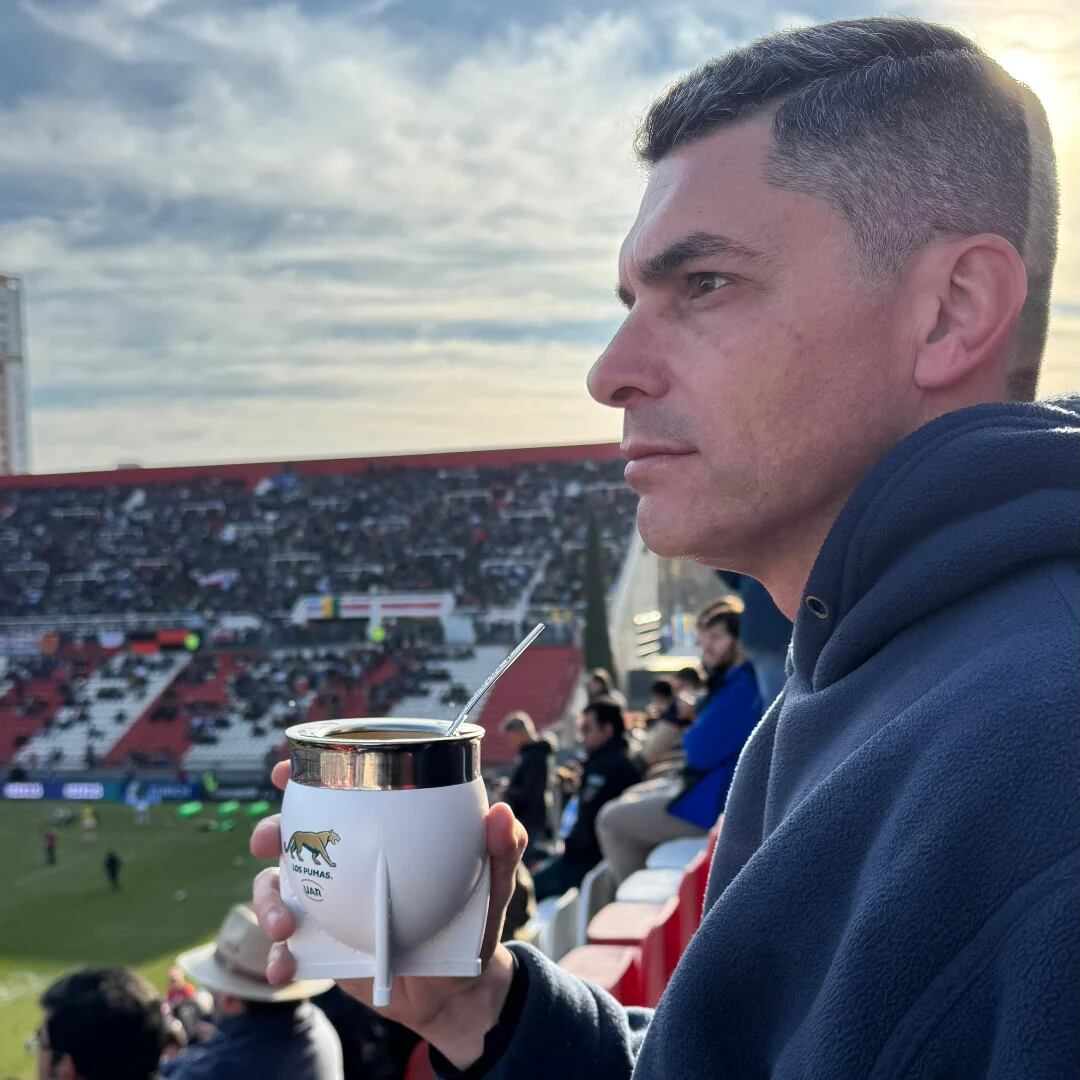 El periodista Santiago Martella sostiene un mate en la tribuna de un estadio durante un encuentro deportivo.