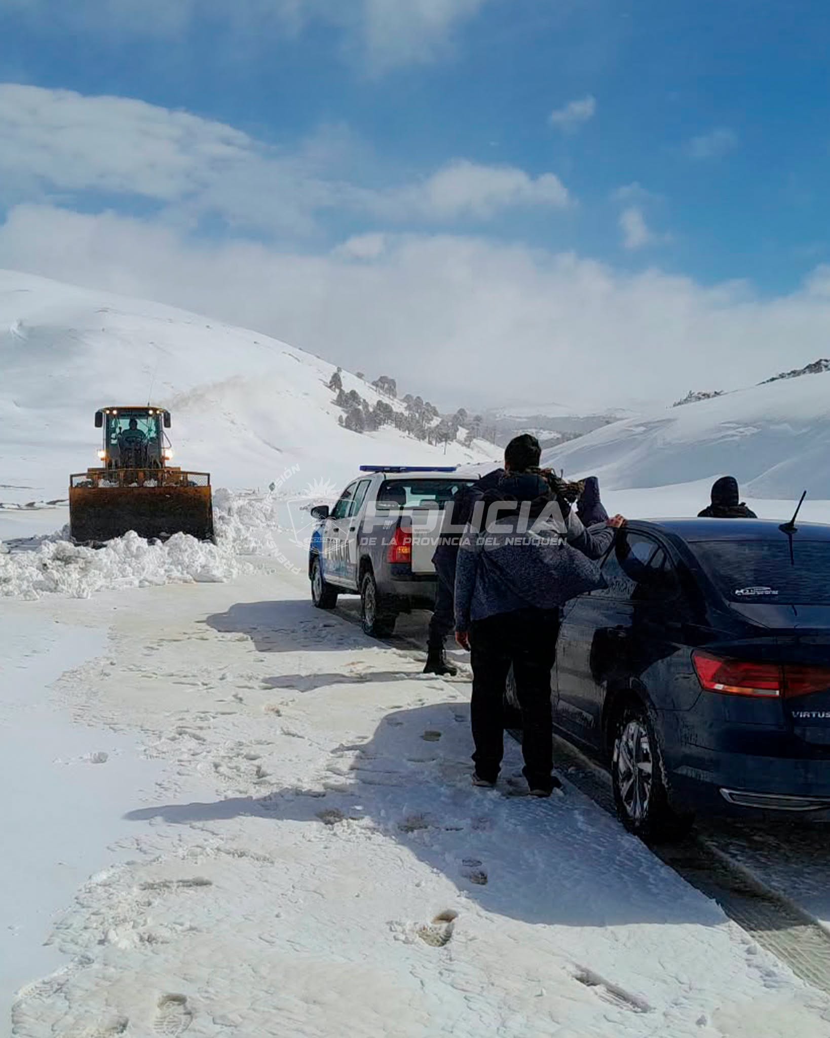 Una máquina vial despeja la nieve de una ruta mientras efectivos de la Policía de Neuquén asisten a los ocupantes de un vehículo en el camino
