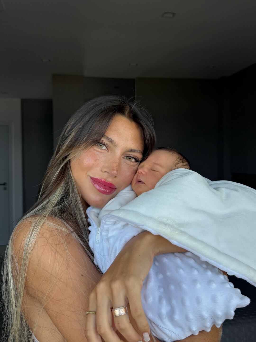 Cielo López, esposa del futbolista Claudio Spinelli, sostiene en sus brazos a su bebé recién nacido envuelto en una manta blanca.
